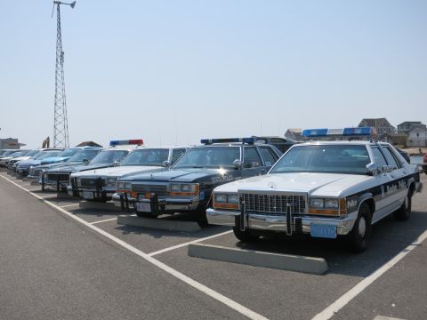 1991 Ford LTD Crown Victoria - Several classic police cruisers lined up in a beachfront lot beside a tall lattice antenna tower.