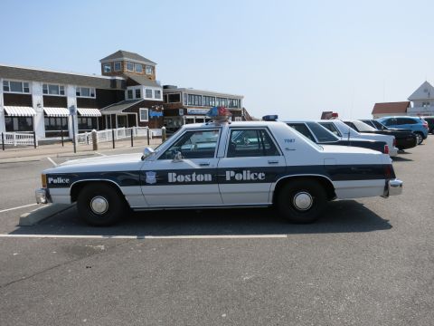 1991 Ford LTD Crown Victoria - black-and-white Boston Police sedan, unit 7087, parked in profile in front of a waterfront building.