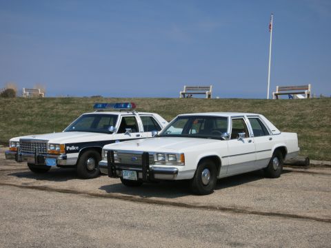 1991 Ford LTD Crown Victoria - blue-and-white police cruiser parked beside an unmarked white Ford LTD Crown Victoria sedan with a flagpole behind.