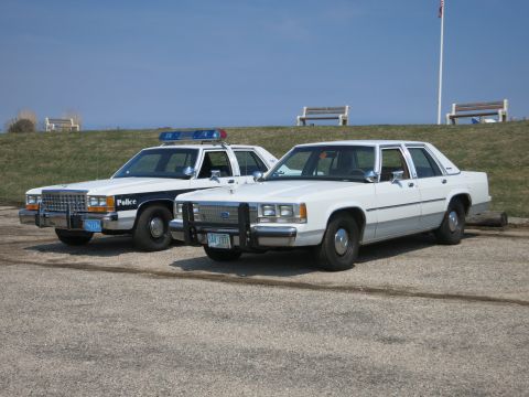 1991 Ford LTD Crown Victoria - marked police cruiser next to an unmarked white Ford sedan in a gravel lot with a grassy embankment behind.