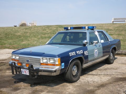 1991 Ford LTD Crown Victoria - two-tone blue State Police Ford sedan, unit 90E, with front push bar parked on a gravel lot.