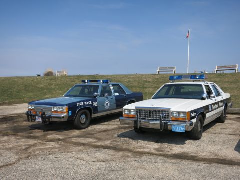 1991 Ford LTD Crown Victoria - blue State Police cruiser parked beside a white Boston Police cruiser on a gravel lot with a flagpole behind.