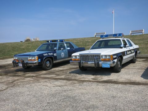 1991 Ford LTD Crown Victoria - blue State Police cruiser and a Boston Police cruiser parked front-angled side by side on a gravel lot.