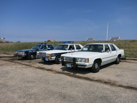 1991 Ford LTD Crown Victoria - Three classic police cruisers, including a State Police and a marked white sedan, parked on a gravel lot.