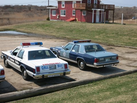 1991 Ford LTD Crown Victoria - Rear view of a Boston Police 'Emergency Call 9-1-1' cruiser and a State Police sedan parked before a red house.