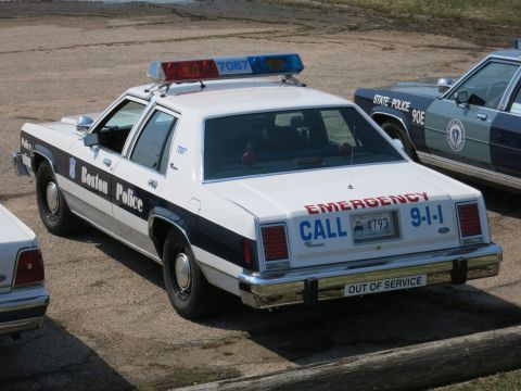 1991 Ford LTD Crown Victoria - Rear quarter view of a Boston Police cruiser marked 'Emergency Call 9-1-1' parked beside a State Police sedan.