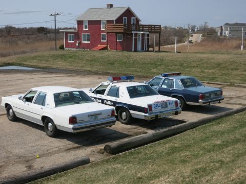 1991 Ford LTD Crown Victoria - Three vintage sedans, including a Boston Police and a State Police cruiser, parked before a red house.