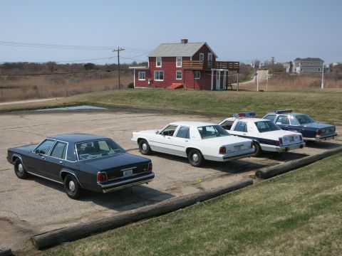 1991 Ford LTD Crown Victoria - Four classic Ford-style sedans, marked and unmarked, parked diagonally on a gravel lot near a red house.