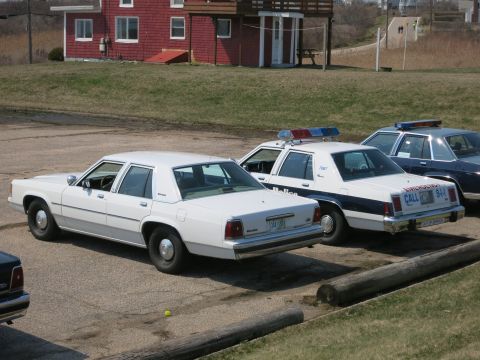 1991 Ford LTD Crown Victoria - white unmarked sedan parked beside marked police cruisers on a gravel lot before a red house.