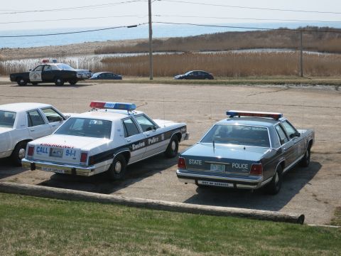 1991 Ford LTD Crown Victoria - Rear view of a Boston Police cruiser and a State Police sedan parked on gravel near the shoreline.