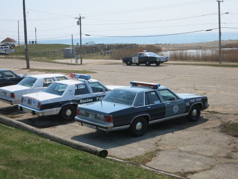 1991 Ford LTD Crown Victoria - lineup of vintage police Ford sedans including Boston Police and State Police cars parked along a coastal lot.