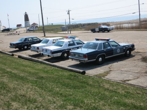 1991 Ford LTD Crown Victoria - Several vintage police Ford sedans, including Boston and State Police cars, parked near a lighthouse by the shore.