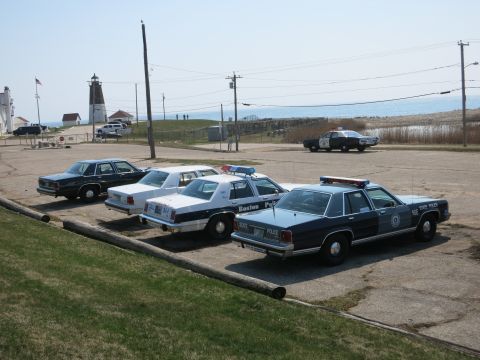 1991 Ford LTD Crown Victoria - row of vintage police Ford sedans parked at a coastal lot near a lighthouse and flagpole.
