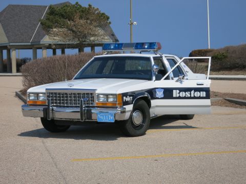 1991 Ford LTD Crown Victoria - Boston Police Ford LTD Crown Victoria sedan with doors open parked on pavement near a pavilion and shrubs.