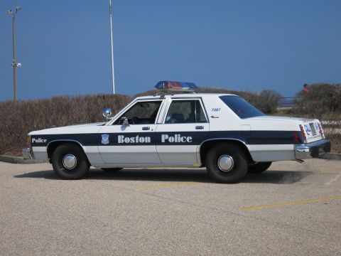 1991 Ford LTD Crown Victoria - Boston Police Ford LTD Crown Victoria sedan in passenger-side profile parked in a lot backed by shrubs.