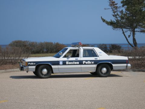 1991 Ford LTD Crown Victoria - Boston Police Ford LTD Crown Victoria sedan in driver-side profile by the shore with windswept trees behind.