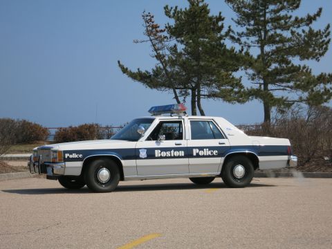 1991 Ford LTD Crown Victoria - Boston Police Ford LTD Crown Victoria sedan in driver-side profile parked at a coastal lot beside pine trees.