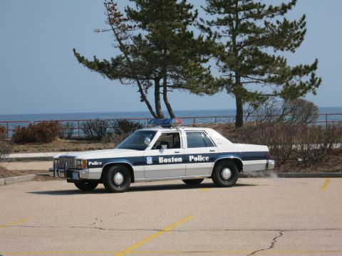 1991 Ford LTD Crown Victoria - Boston Police Ford LTD Crown Victoria sedan in driver-side profile beside windswept trees overlooking the ocean.
