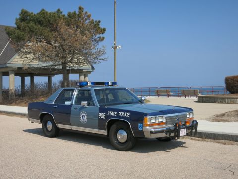 1991 Ford LTD Crown Victoria - two-tone blue and silver Massachusetts State Police sedan marked 90E sits on pavement near a beach pavilion under clear skies.