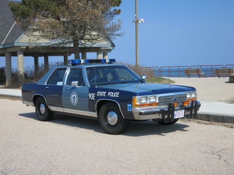 1991 Ford LTD Crown Victoria - Front three-quarter view of a blue and silver State Police sedan with push bar and 90E markings, parked beside a coastal railing.