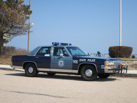 1991 Ford LTD Crown Victoria - Side profile of a two-tone blue and silver State Police sedan with light bar, parked on a coastal road near a railing.