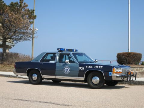 1991 Ford LTD Crown Victoria - blue and silver Massachusetts State Police sedan marked 90E with a front push bar, parked along a seaside road.