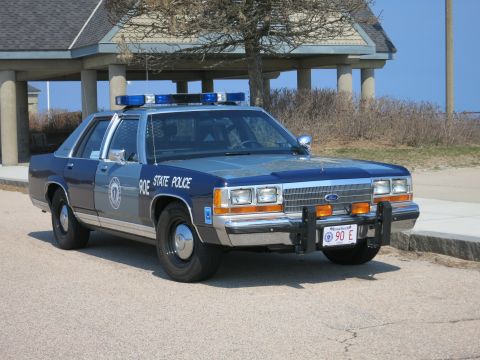 1991 Ford LTD Crown Victoria - Front view of a blue and silver Ford State Police sedan marked 90E, with chrome grille, push bar, and roof lights near a pavilion.