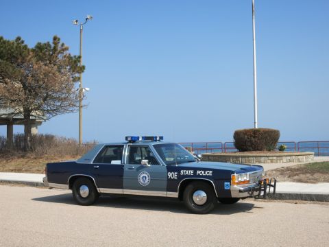 1991 Ford LTD Crown Victoria - blue and silver Massachusetts State Police sedan marked 90E parked on a paved road beside the ocean and a railing.