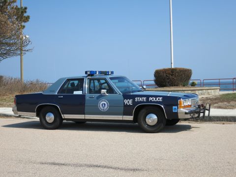 1991 Ford LTD Crown Victoria - blue and silver Massachusetts State Police sedan marked 90E sits on a coastal road with a flagpole and shrubs nearby.