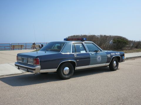 1991 Ford LTD Crown Victoria - Rear three-quarter view of a two-tone blue and silver State Police sedan parked along a seaside walkway with shrubs.