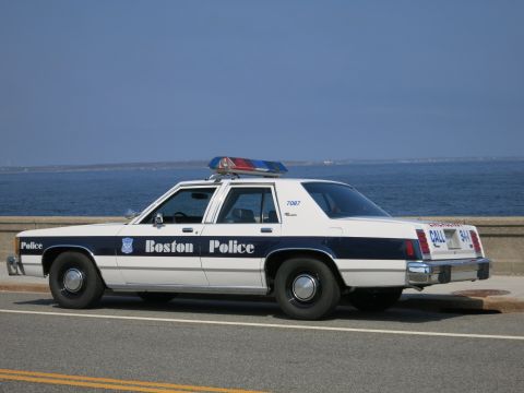 1991 Ford LTD Crown Victoria - white and blue Boston Police Ford sedan marked 7087 with EMERGENCY CALL 911 lettering, parked on a road beside the ocean.