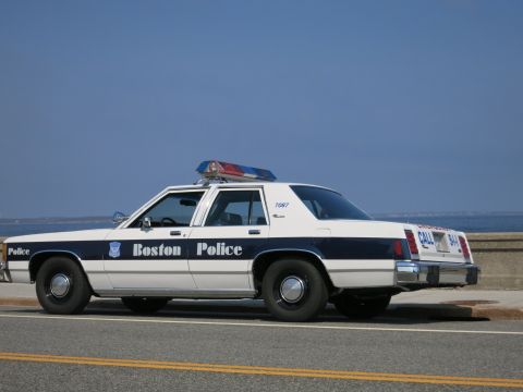 1991 Ford LTD Crown Victoria - Rear three-quarter view of a white and blue Boston Police Ford sedan marked 7087 with light bar, parked beside the sea.