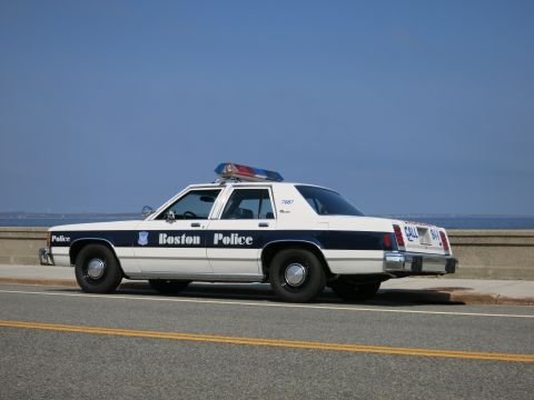 1991 Ford LTD Crown Victoria - white and blue Boston Police sedan with roof light bar parked along a seawall road under a clear blue sky.