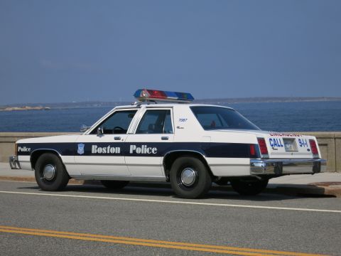 1991 Ford LTD Crown Victoria - Rear three-quarter view of a white and blue Boston Police sedan marked 7087 with CALL 911 lettering, parked by the sea.