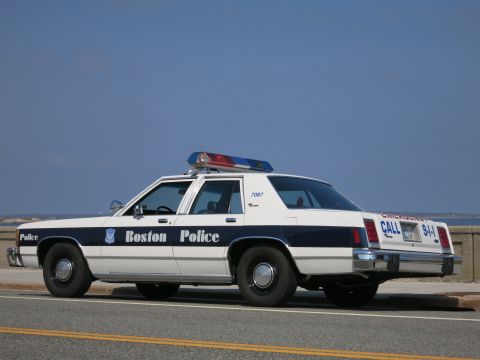 1991 Ford LTD Crown Victoria - Rear three-quarter view of a white and blue Boston Police Ford sedan marked 7087 with red and blue roof lights, beside the ocean.