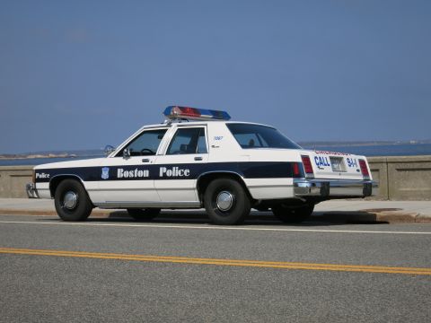 1991 Ford LTD Crown Victoria - white and blue Boston Police sedan marked 7087 with EMERGENCY CALL 911 graphics parked along a coastal road.