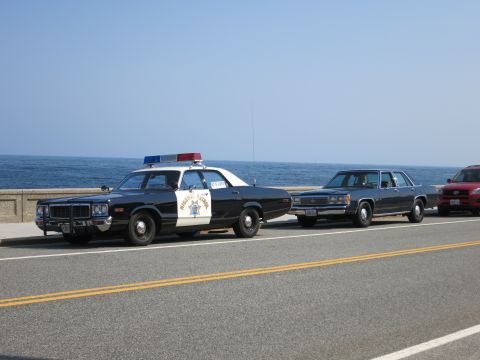 1991 Ford LTD Crown Victoria - black and white Highway Patrol sedan with light bar parked on a coastal road ahead of a dark sedan and a red car.