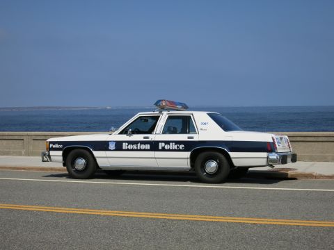 1991 Ford LTD Crown Victoria - Side profile of a white and blue Boston Police sedan marked 7087 with roof light bar, parked beside the ocean.
