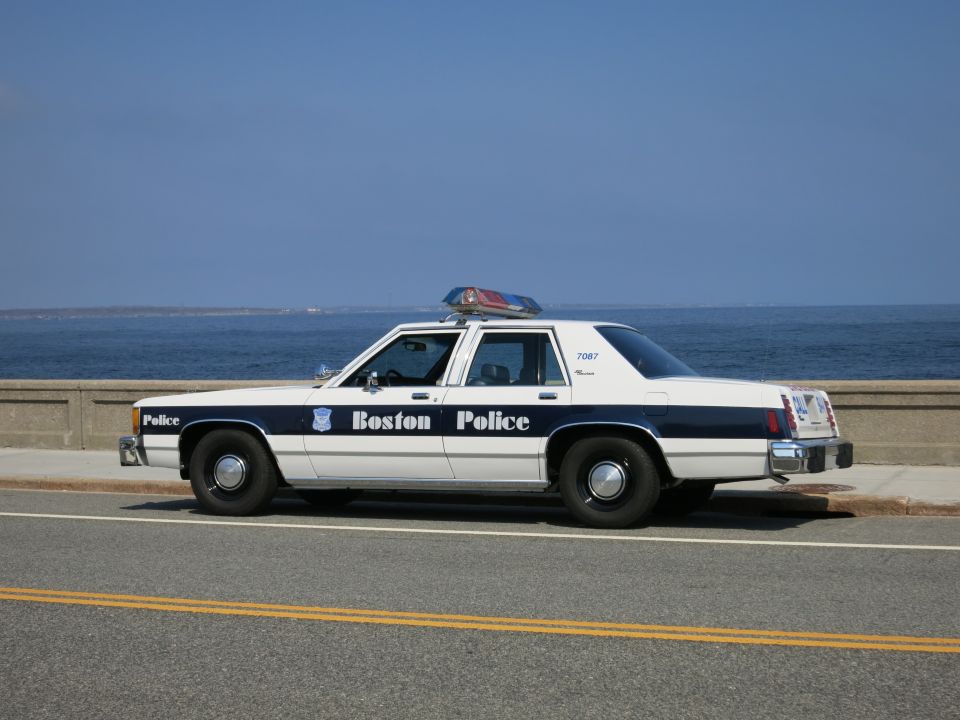 Side view of a white and blue Boston Police sedan along a seawall