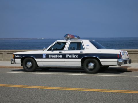 1991 Ford LTD Crown Victoria - Driver-side profile of a white and blue Boston Police sedan marked 7087, parked along a seawall under clear skies.