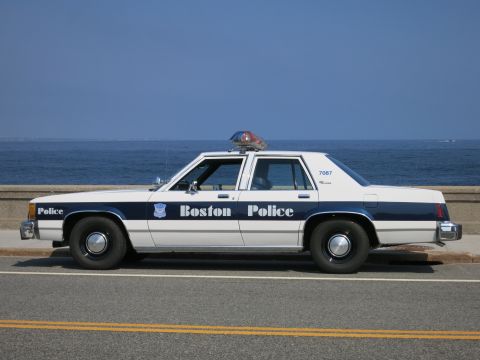 1991 Ford LTD Crown Victoria - Side view of a white and blue Boston Police sedan marked 7087 with roof light bar, parked beside a seawall and the ocean.