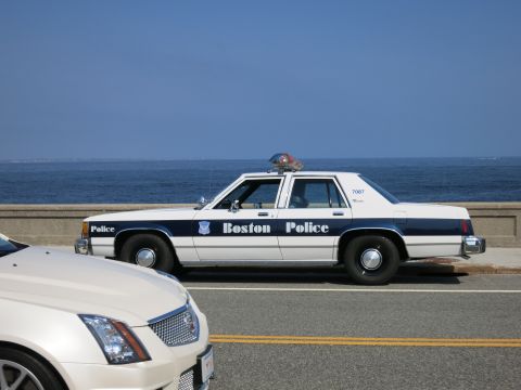 1991 Ford LTD Crown Victoria - white and blue Boston Police sedan marked 7087 parked along a seawall, with a white luxury coupe in the foreground.