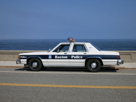 1991 Ford LTD Crown Victoria - Side profile of a white and blue Boston Police sedan marked 7087 with roof light bar, parked beside the ocean.