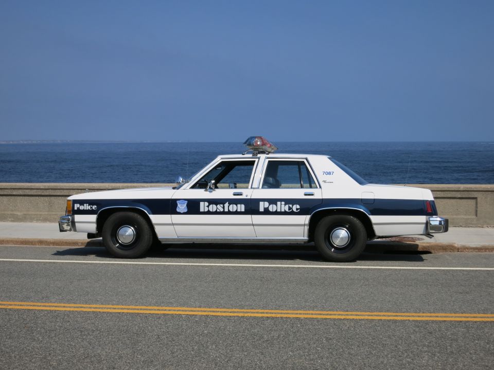 Side profile of a white and blue Boston Police sedan along the coast
