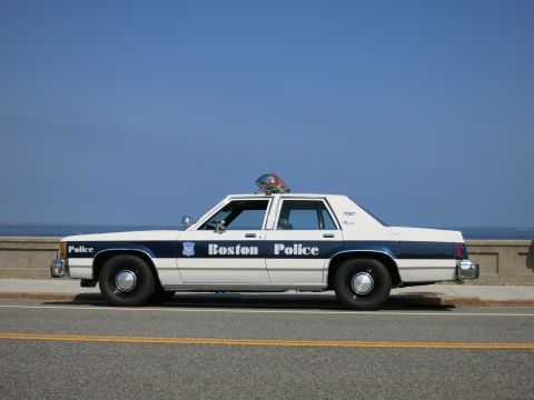 1991 Ford LTD Crown Victoria - Driver-side view of a white and blue Boston Police sedan marked 7087 with roof light bar, parked along a coastal road.