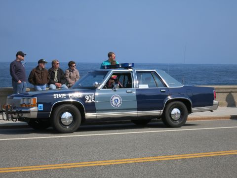 1991 Ford LTD Crown Victoria - Several people sit on the hood of a blue State Police sedan marked 90E parked along a seawall by the ocean.