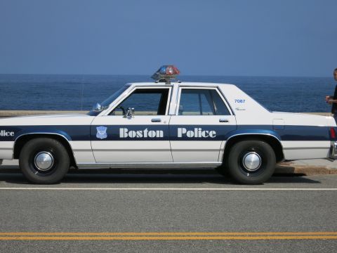 1991 Ford LTD Crown Victoria - Driver-side view of a white-and-blue Boston Police sedan, unit 7087, parked beside a coastal seawall.
