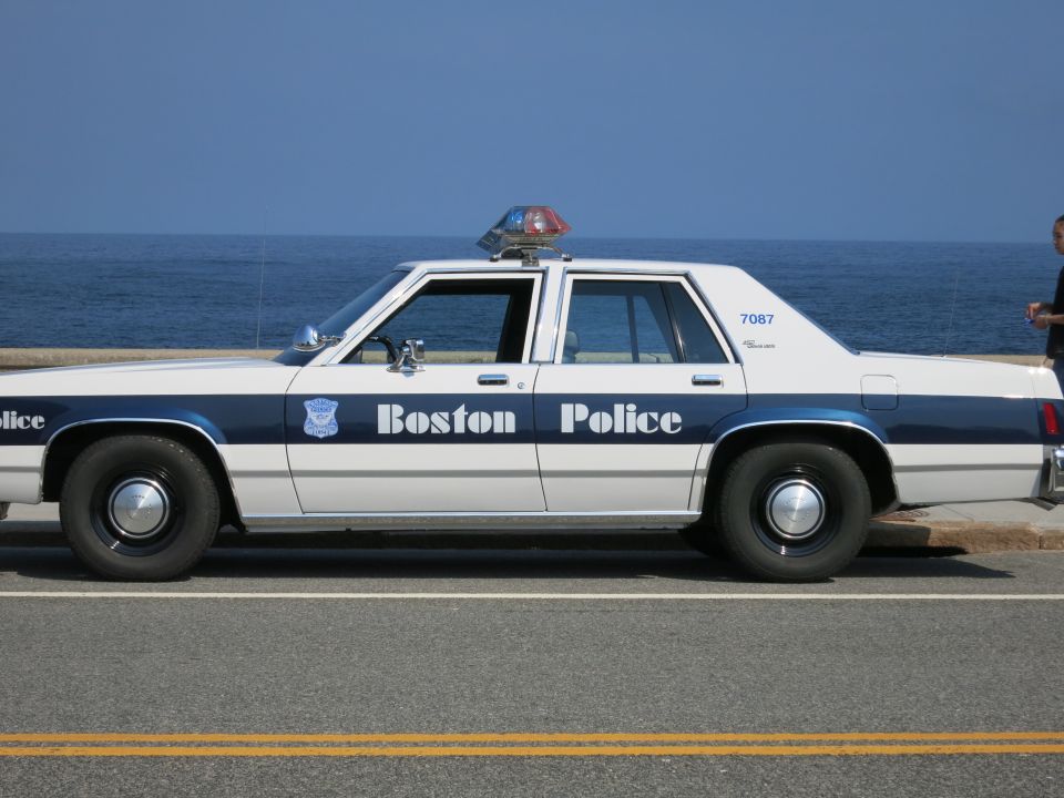 Side view of a white and blue Boston Police sedan parked by the ocean