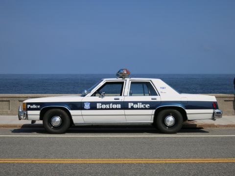 1991 Ford LTD Crown Victoria - Full side profile of a white-and-blue Boston Police Ford sedan, unit 7087, parked on a road beside the ocean.