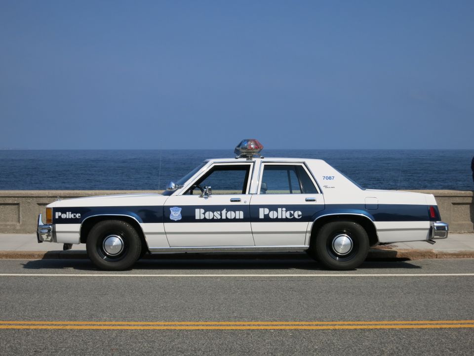 Profile of a white and blue Boston Police sedan along a seawall road